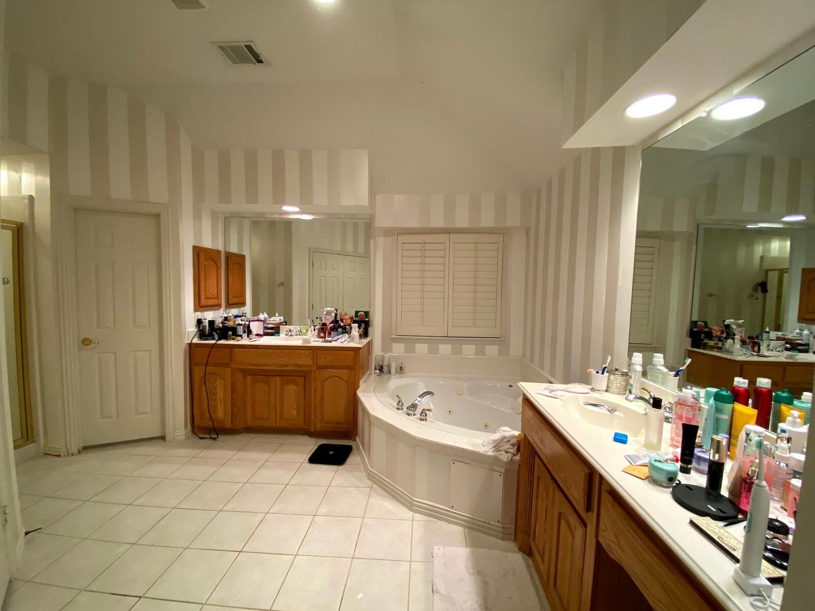 Before: primary bath with striped wallpaper, oak vanities, and a tiled corner jetted tub