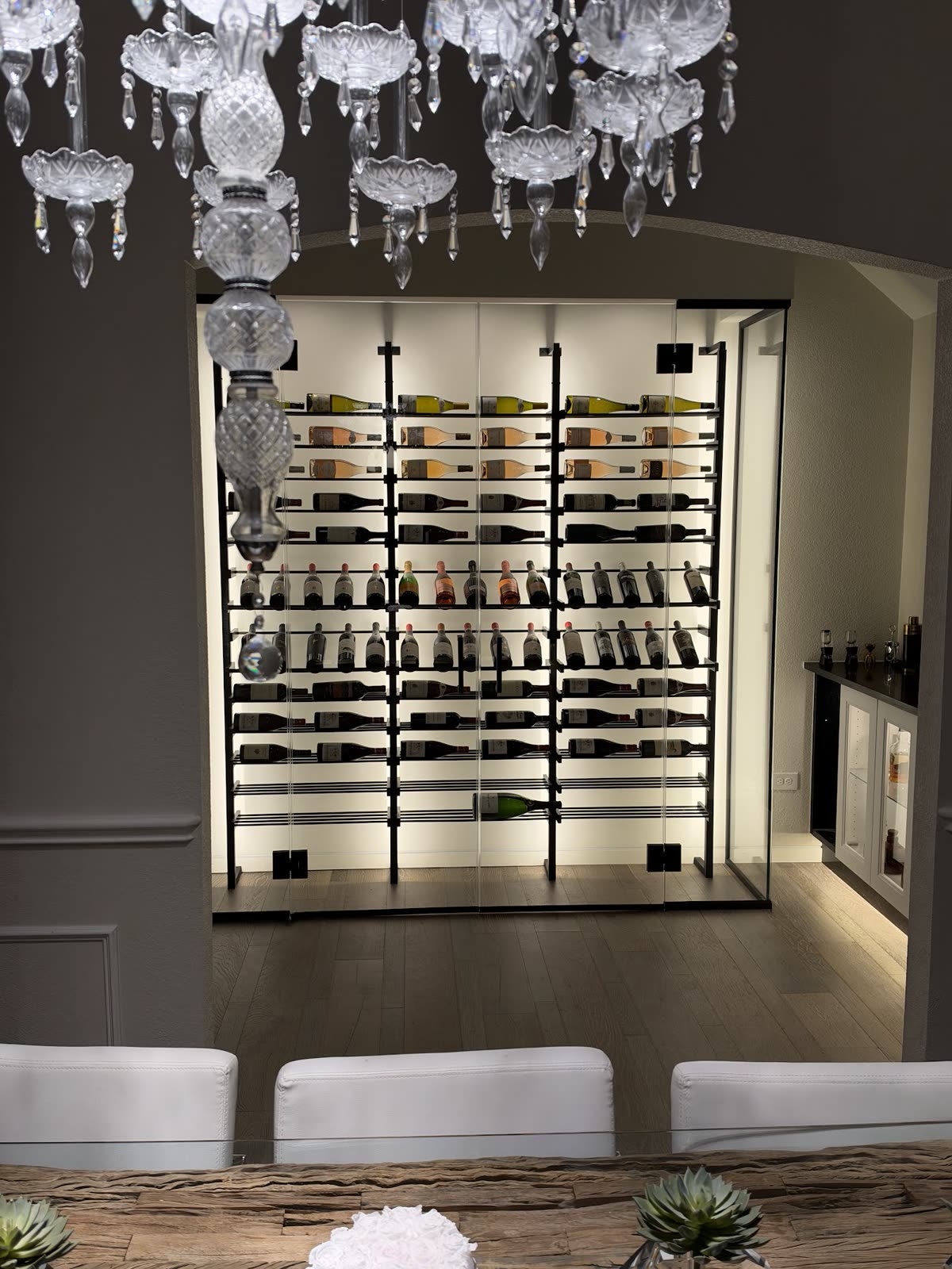 Finished glass wine room, stocked and lit, seen through an arched opening from the dining room under a crystal chandelier, with the bar to the right