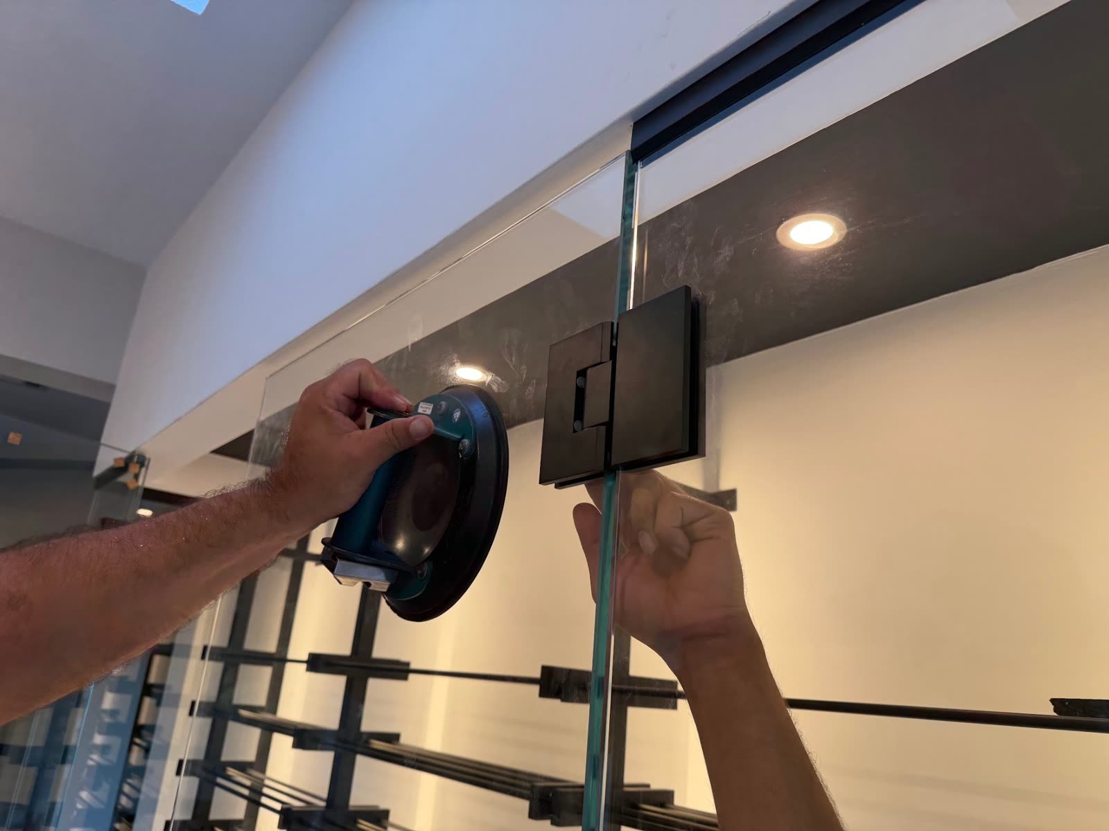 Hands holding a glass panel with a suction-cup lifter while a black glass-door hinge is set, with the steel racking behind the glass